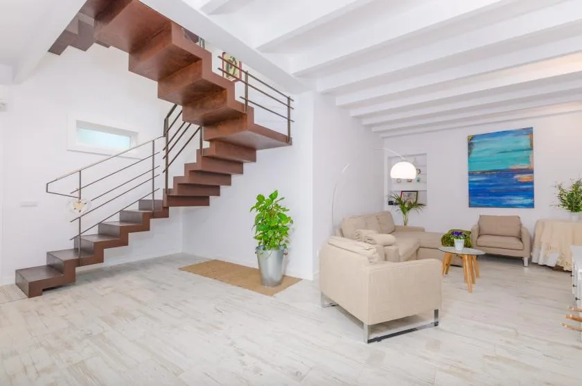 A bright living room with seating, a staircase, and a colorful wall painting.