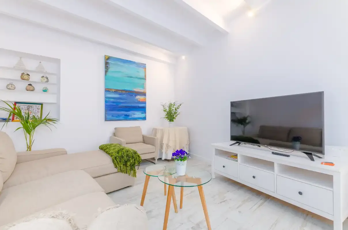 A bright living room with light seating, a glass coffee table, and a television.