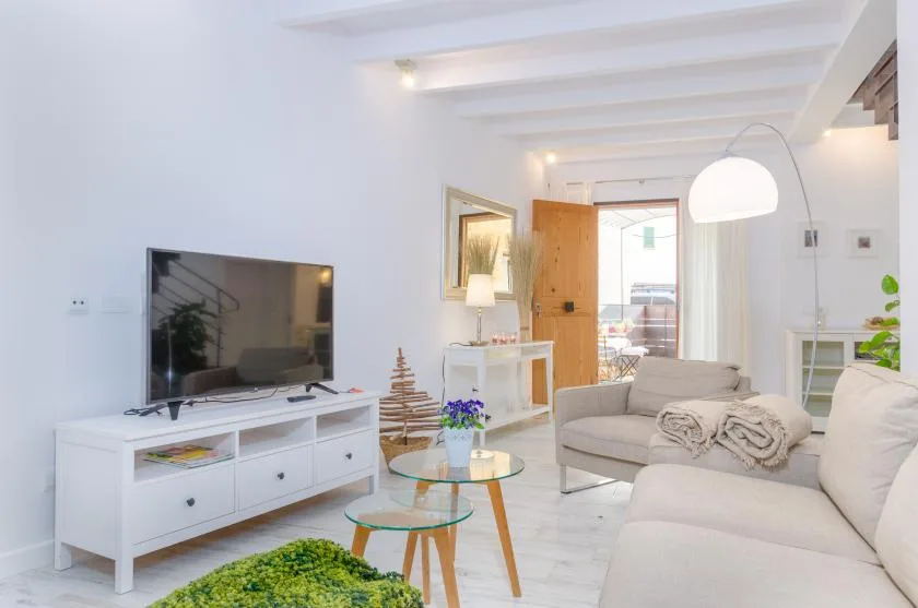 A bright living room with pale seating, a television, and a glass coffee table.