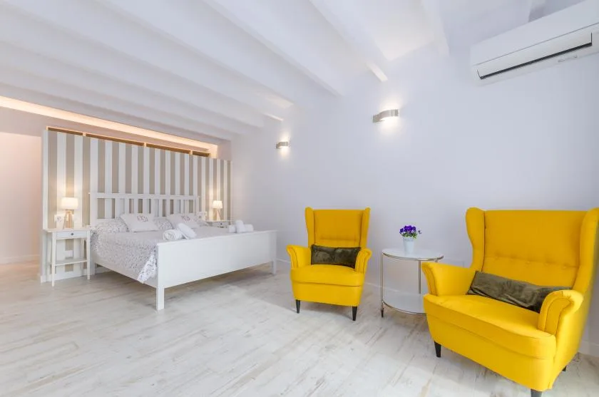 A bright bedroom with a white bed and yellow upholstered seating.