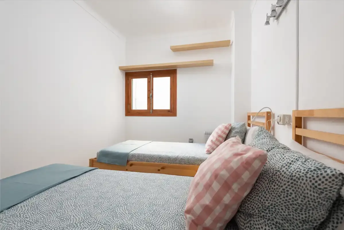 A bright bedroom with two beds and a small wooden-framed window.