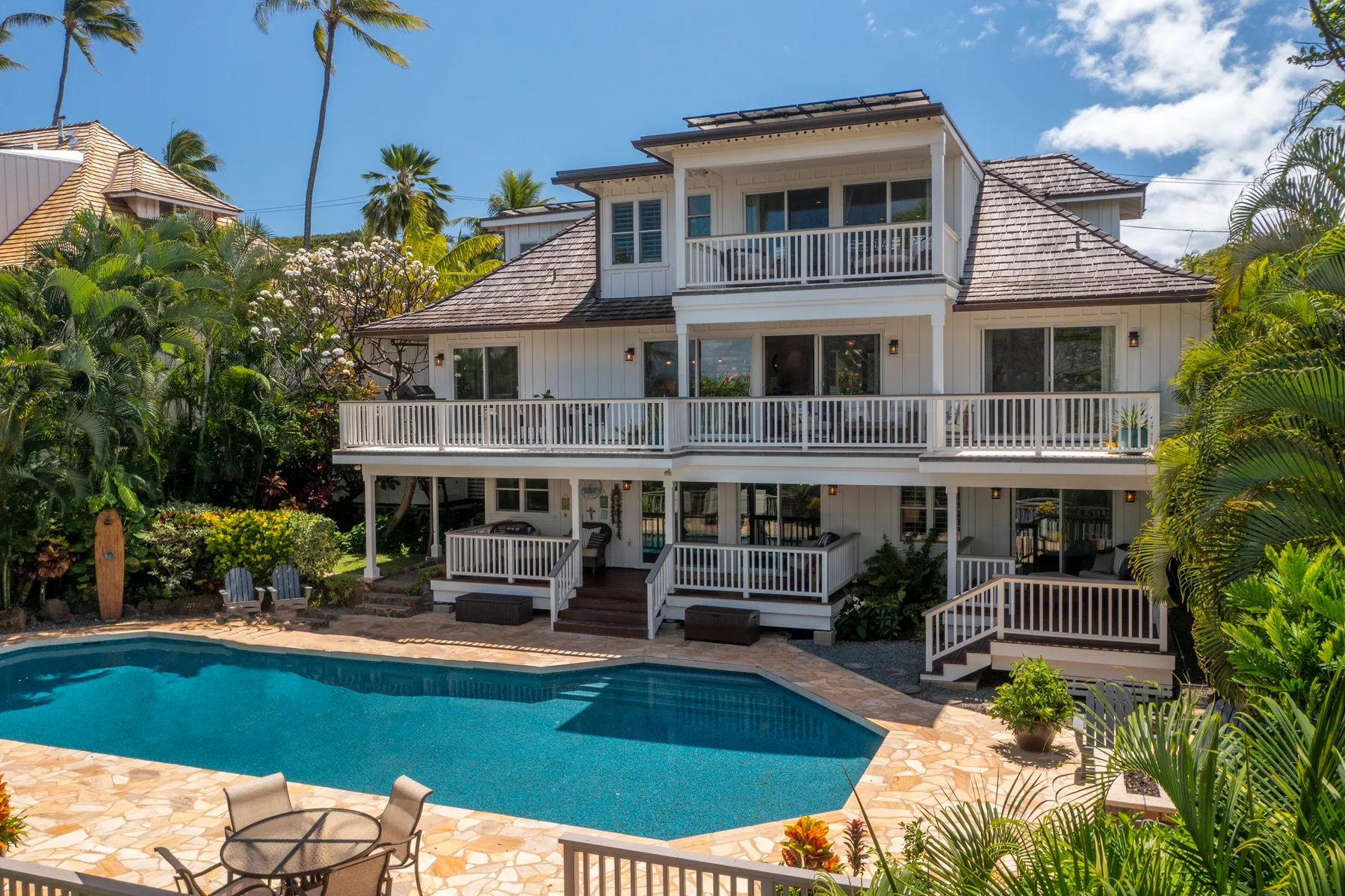 A large white house overlooks a swimming pool surrounded by tropical greenery.