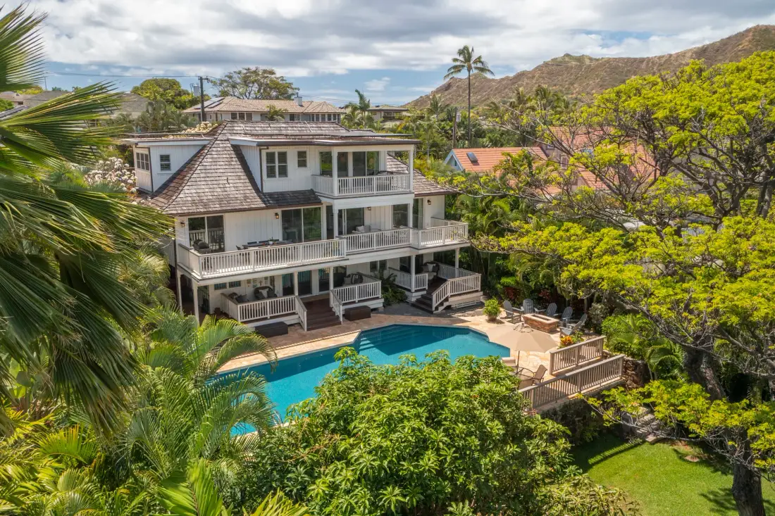 A multi-level house overlooks a swimming pool amid lush trees.