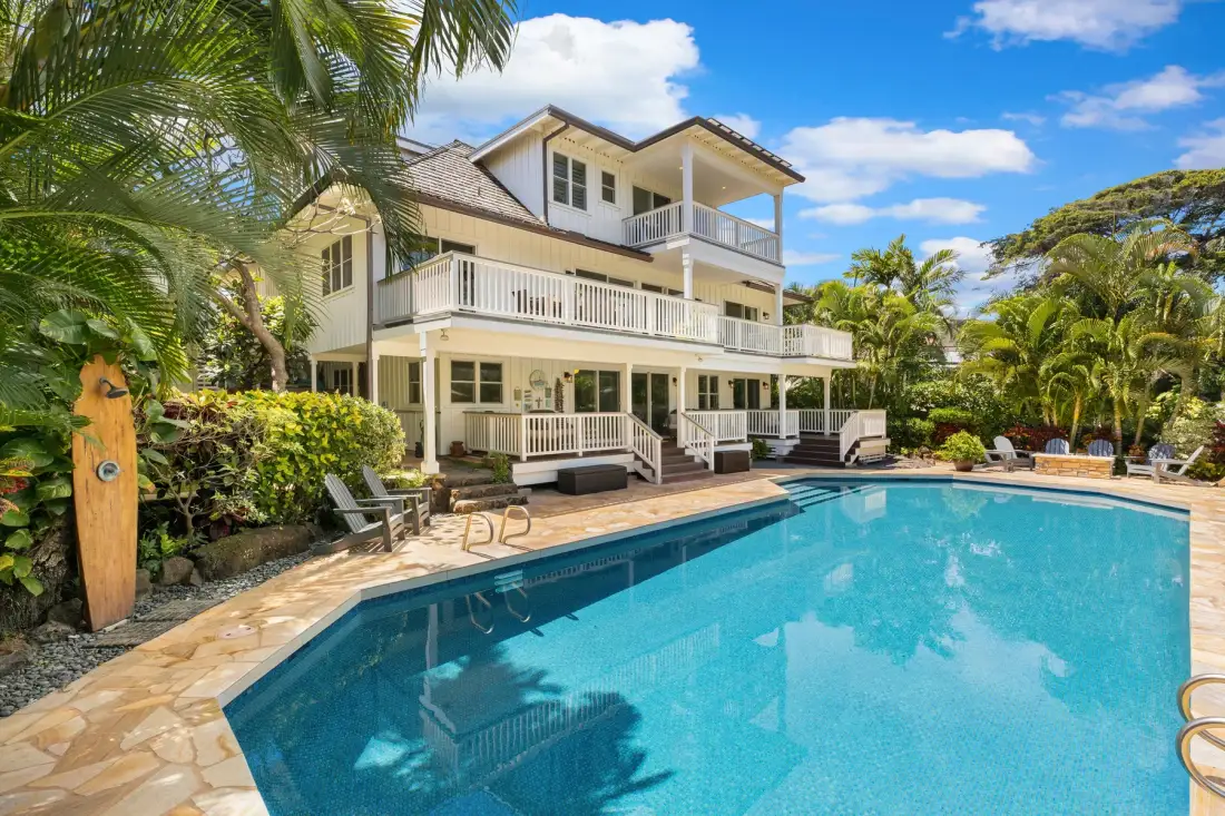 A large outdoor swimming pool sits beside a multi-story house and surrounding tropical greenery.