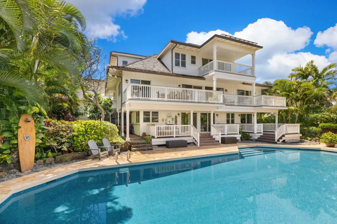 A large white house overlooks a swimming pool surrounded by tropical landscaping.