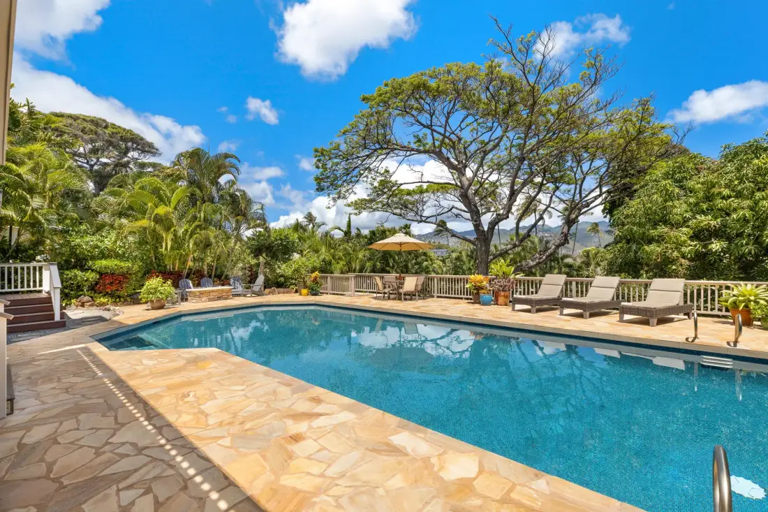 An outdoor swimming pool is surrounded by a stone deck, seating, and leafy trees.