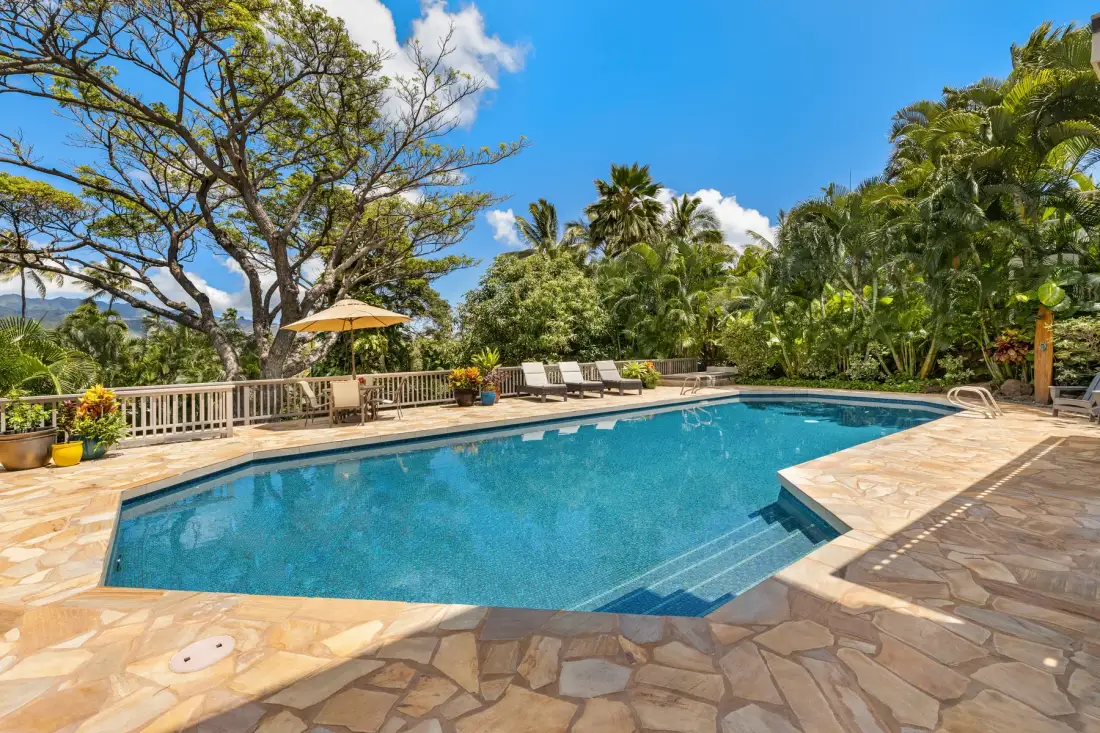 A swimming pool sits beside a paved patio, lounge seating, and tropical greenery.