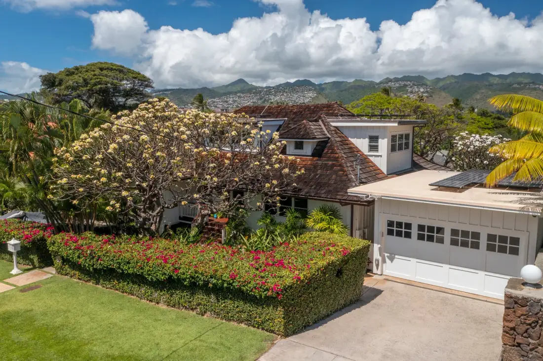 A house and garage stand beside a landscaped garden, with hills in the distance.