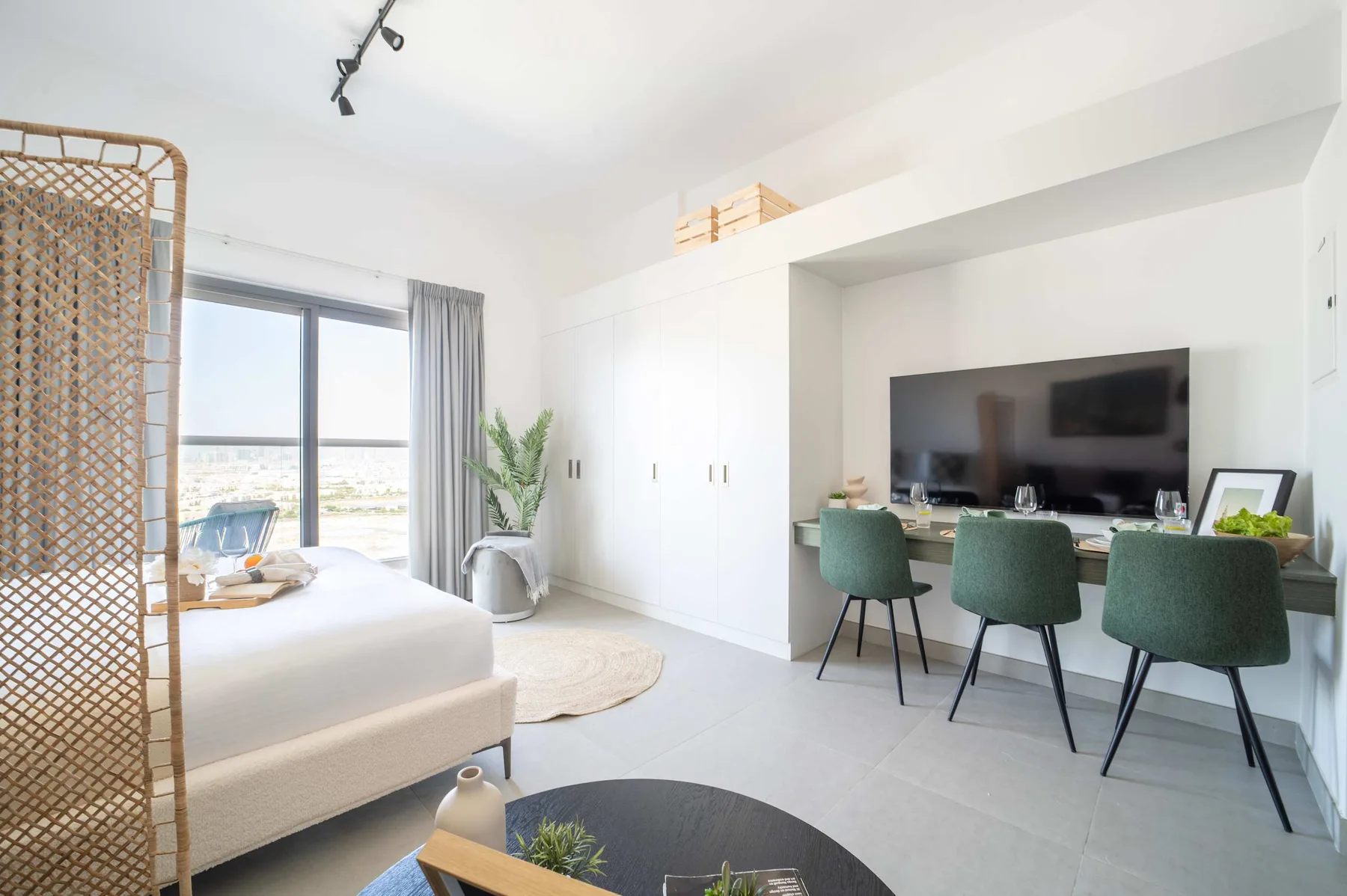 Un dormitorio luminoso con una cama junto a un escritorio empotrado con televisión y asientos.