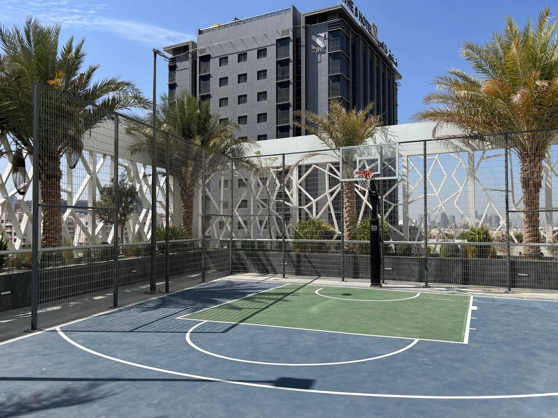 An outdoor basketball court with a hoop, palm trees, and a tall building in the background.