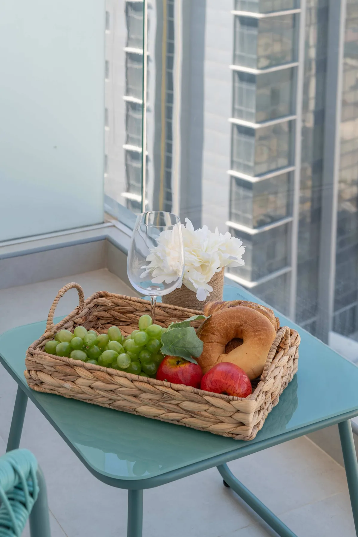 A glass table by a window holds a basket of fruit and a vase of flowers.