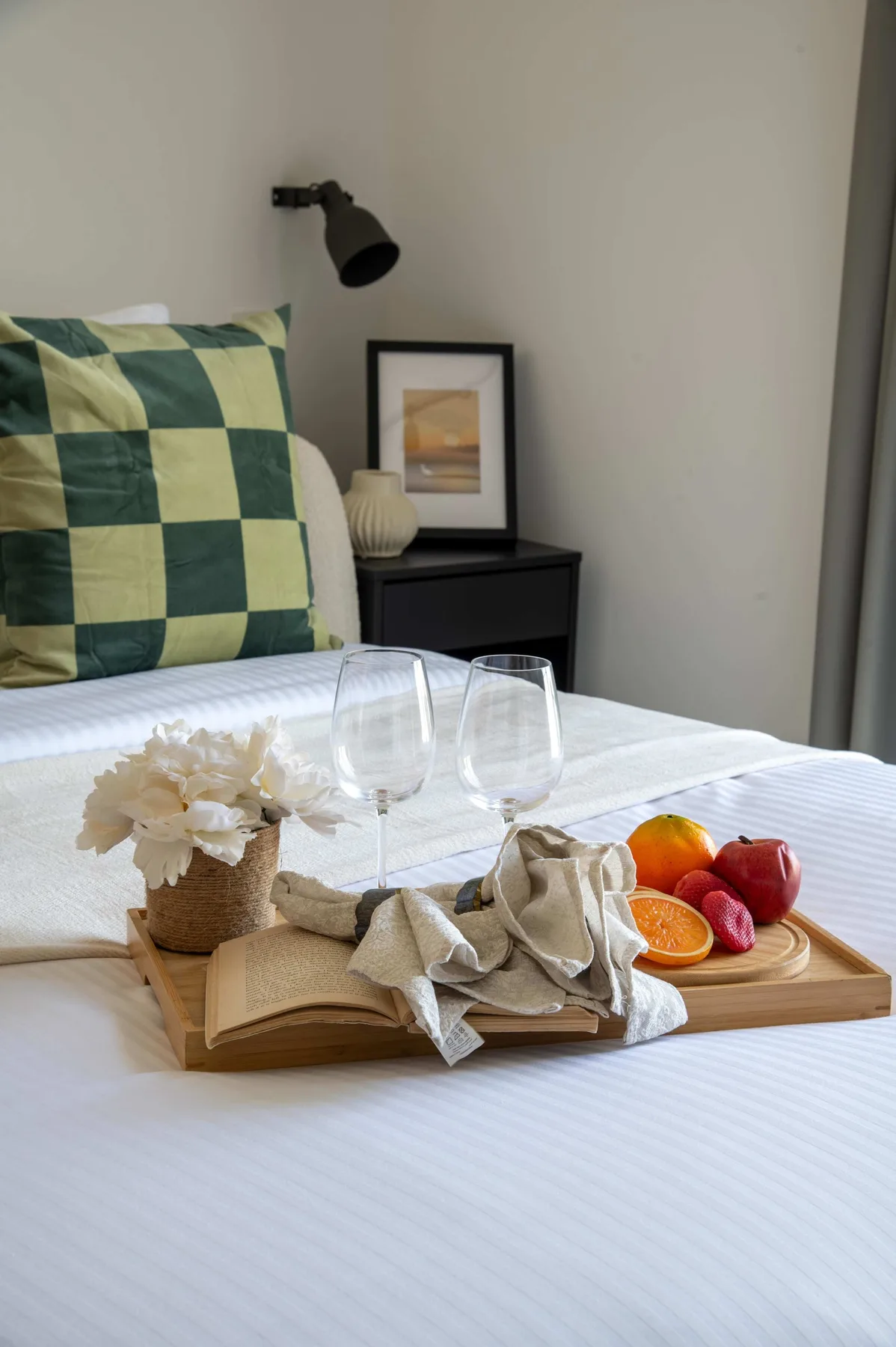 A neatly made bedroom features a tray with glasses, flowers, and fruit on the bed.