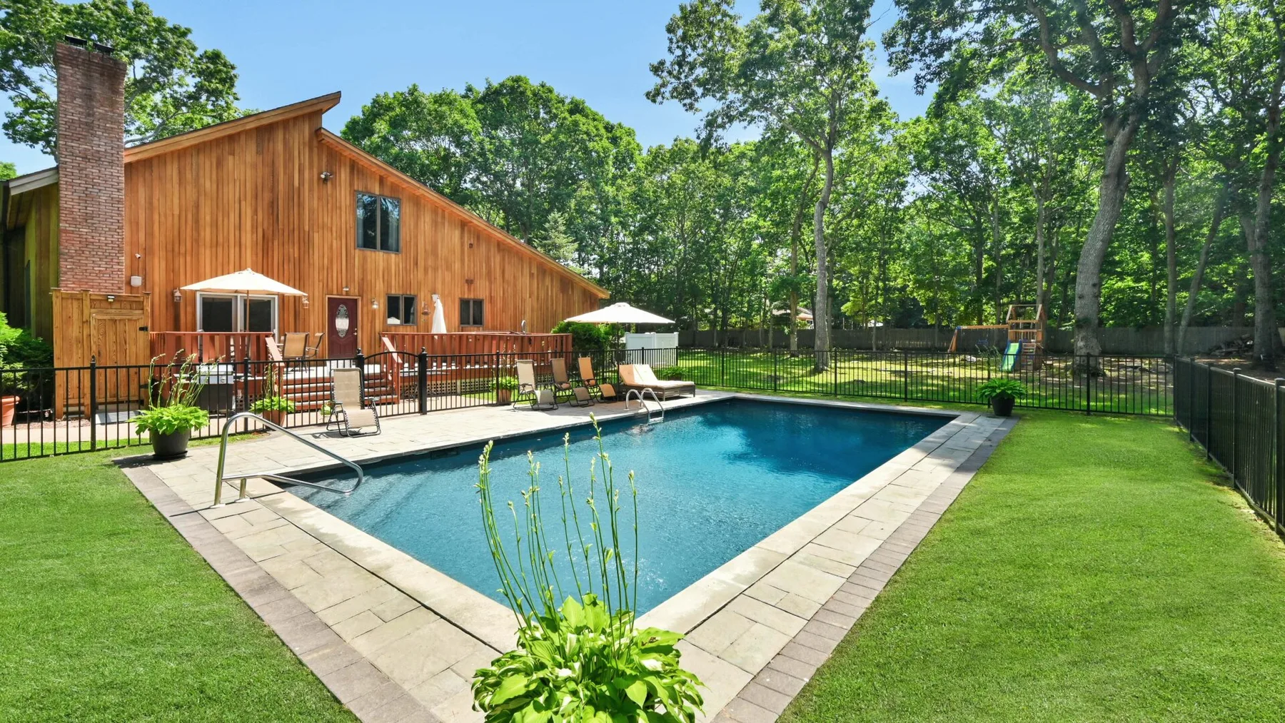Una piscina está junto a una casa con revestimiento de madera, asientos de patio y césped verde.