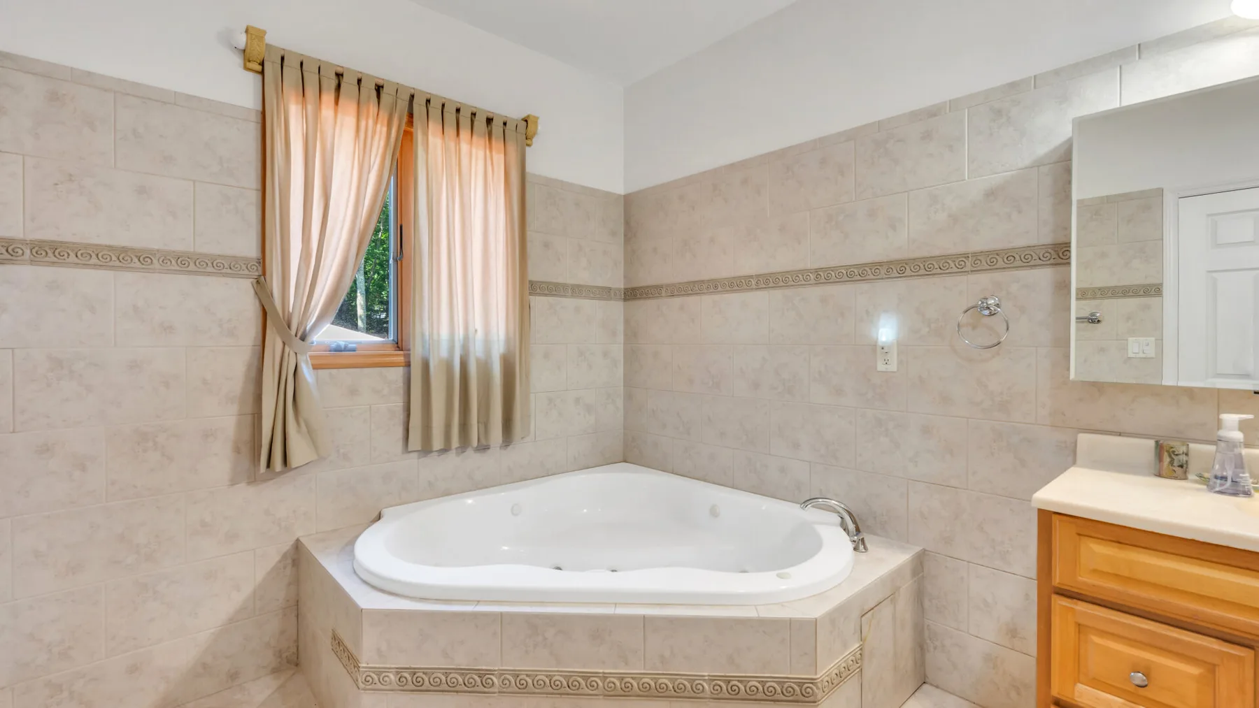 Baño alicatado con bañera empotrada, ventana con cortinas y mueble de madera.