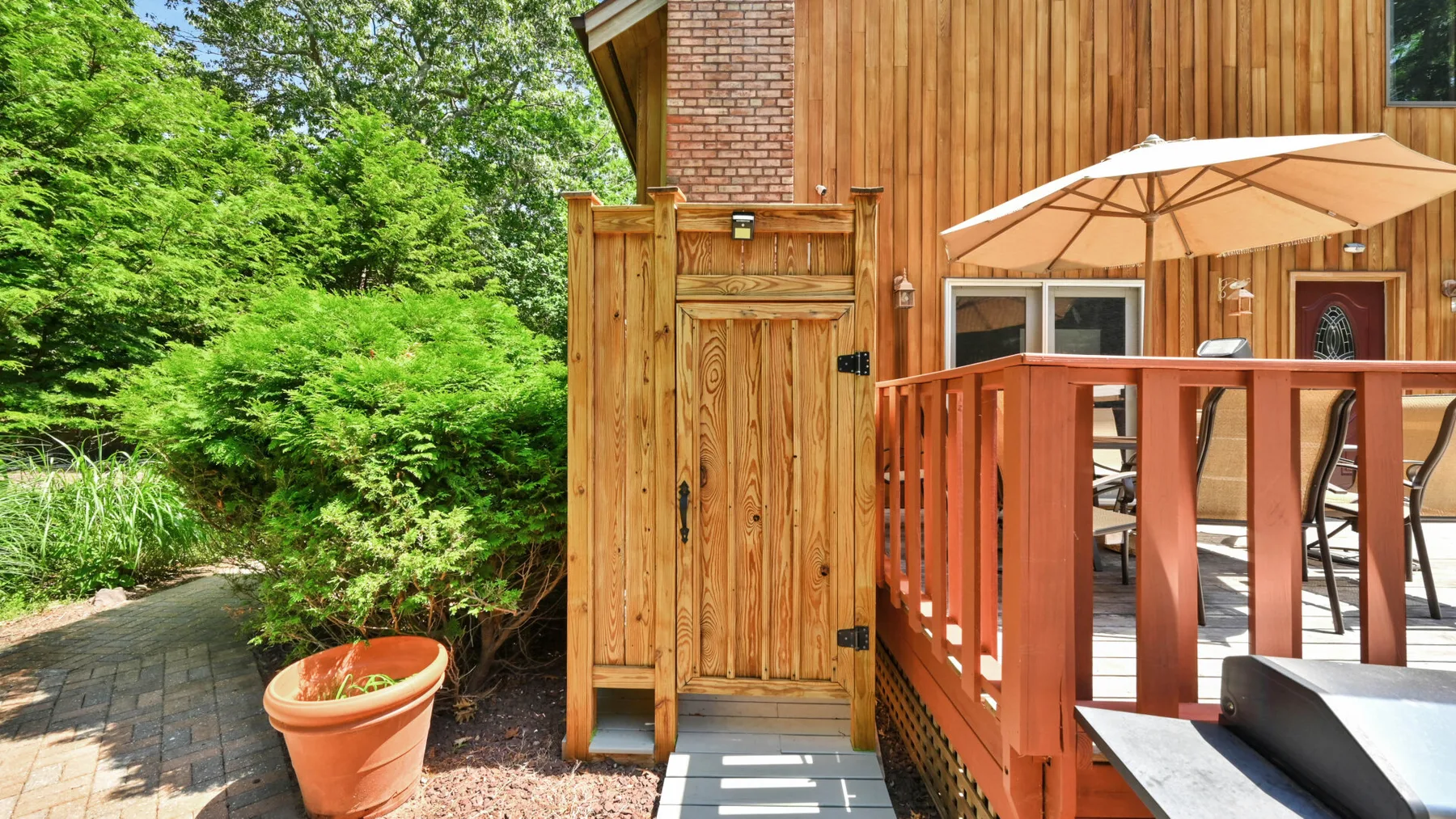 Exterior de una cabaña de madera con una pequeña terraza, sombrilla y vegetación alrededor.