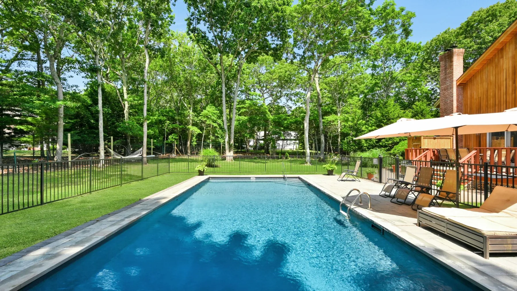 Una piscina al aire libre junto a una terraza de madera y un césped arbolado.