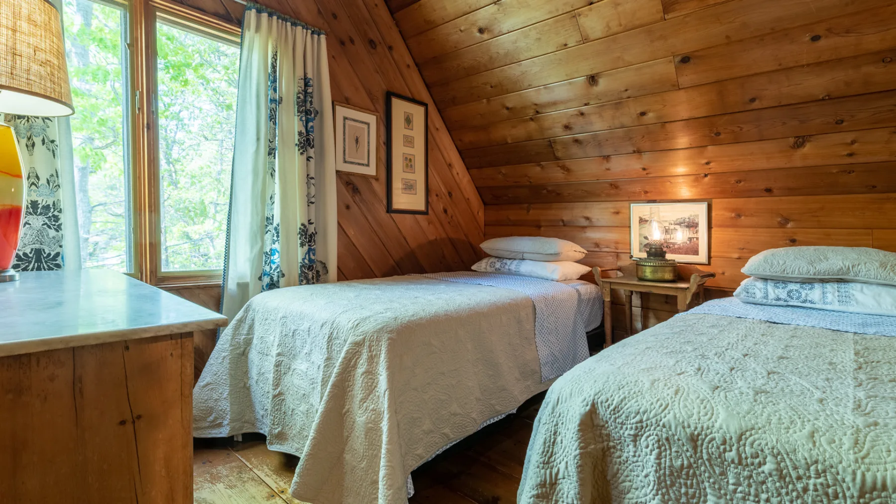 Dormitorio revestido de madera con dos camas y una ventana con vistas a la vegetación.