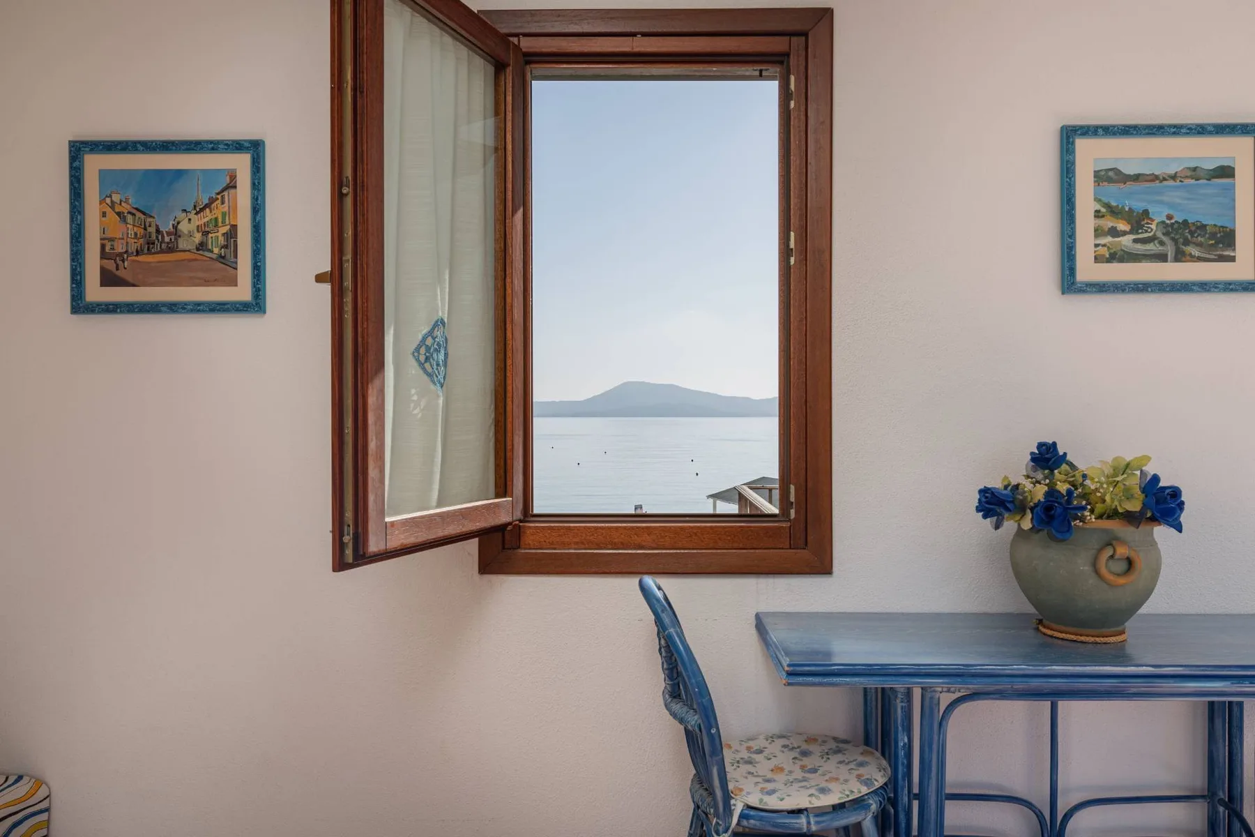 Una habitación luminosa tiene una ventana abierta con vistas al mar y una mesa cerca.