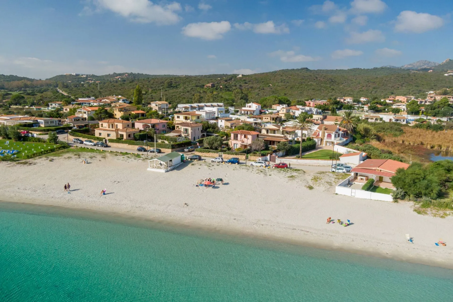 Una playa de arena bordea aguas turquesas, con casas y colinas verdes al fondo.