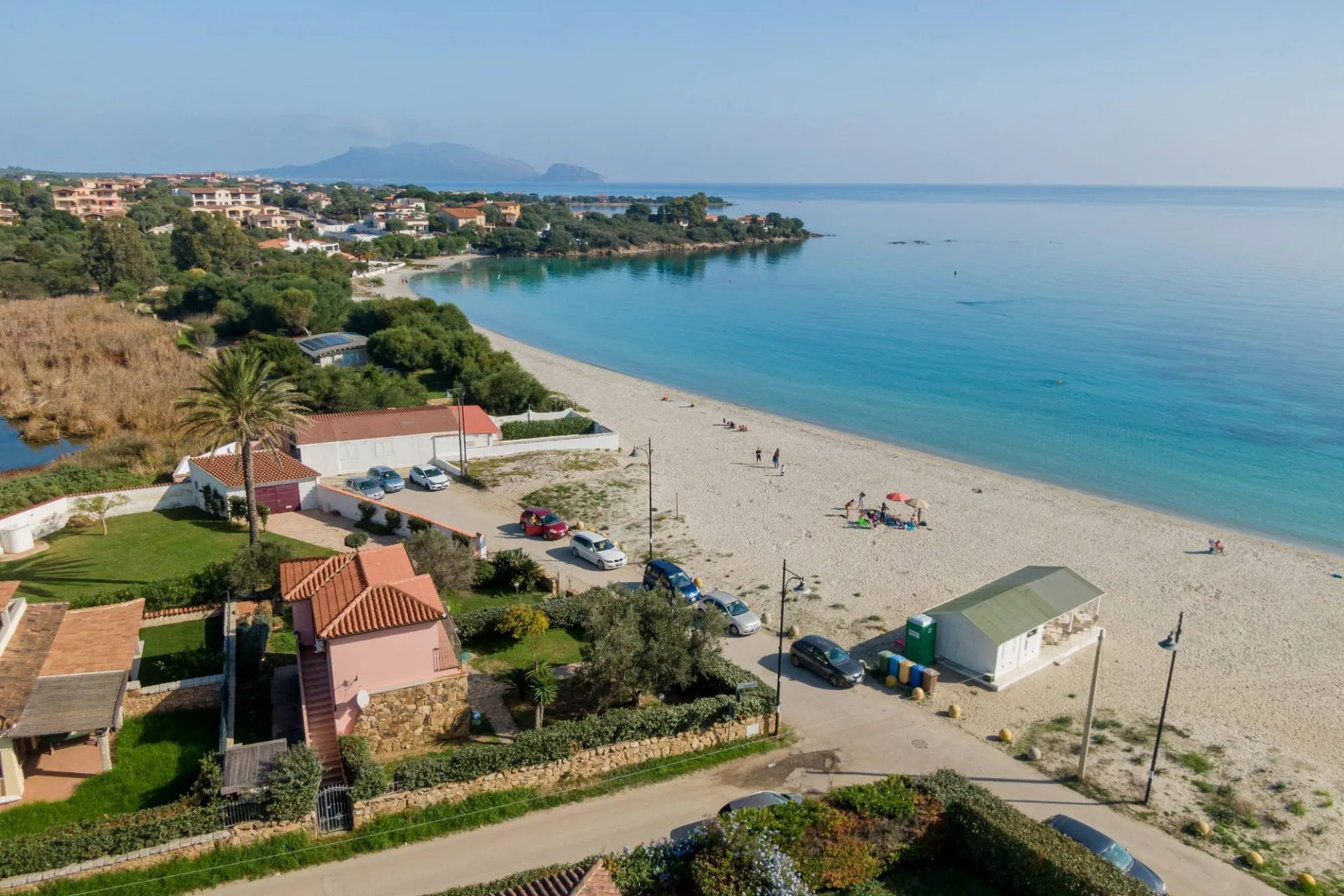 Una playa de arena bordea aguas azules y tranquilas, junto a casas y vegetación costeras.