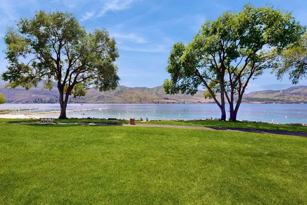 A grassy lakeside lawn with trees framing the water and distant hills.
