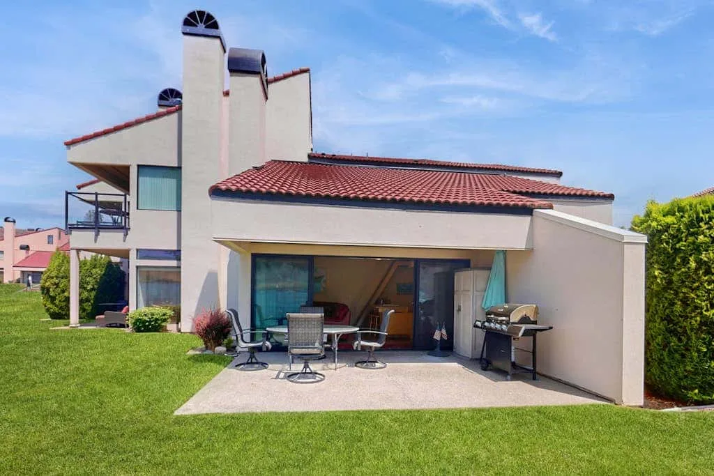 A modern house opens onto a patio with outdoor seating and a grill.