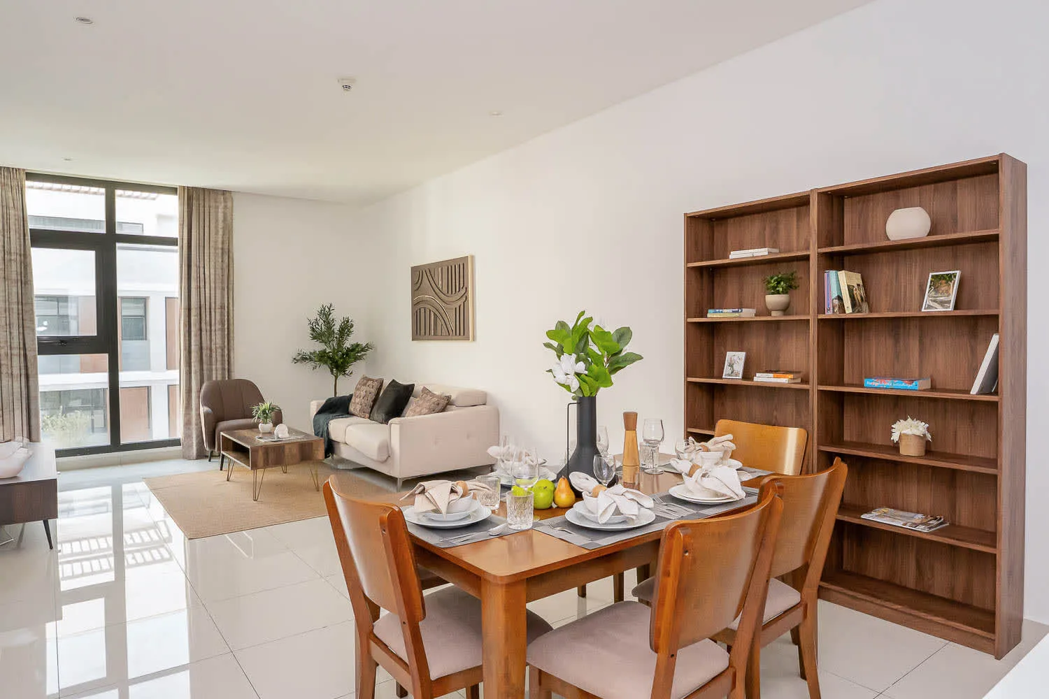 A bright dining and living area features a set dining table beside a tall wooden bookcase.