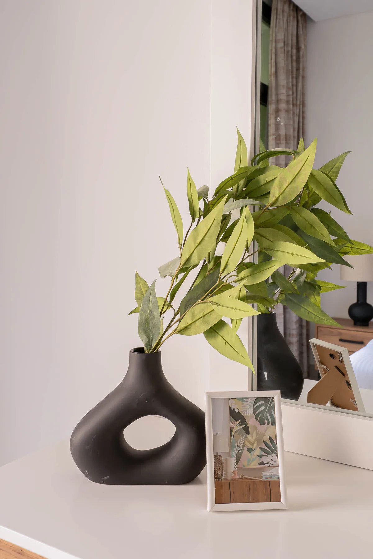 A framed botanical print and leafy branches sit on a white bedroom dresser.