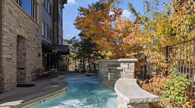 An outdoor swimming pool curves beside a stone building and leafy trees.