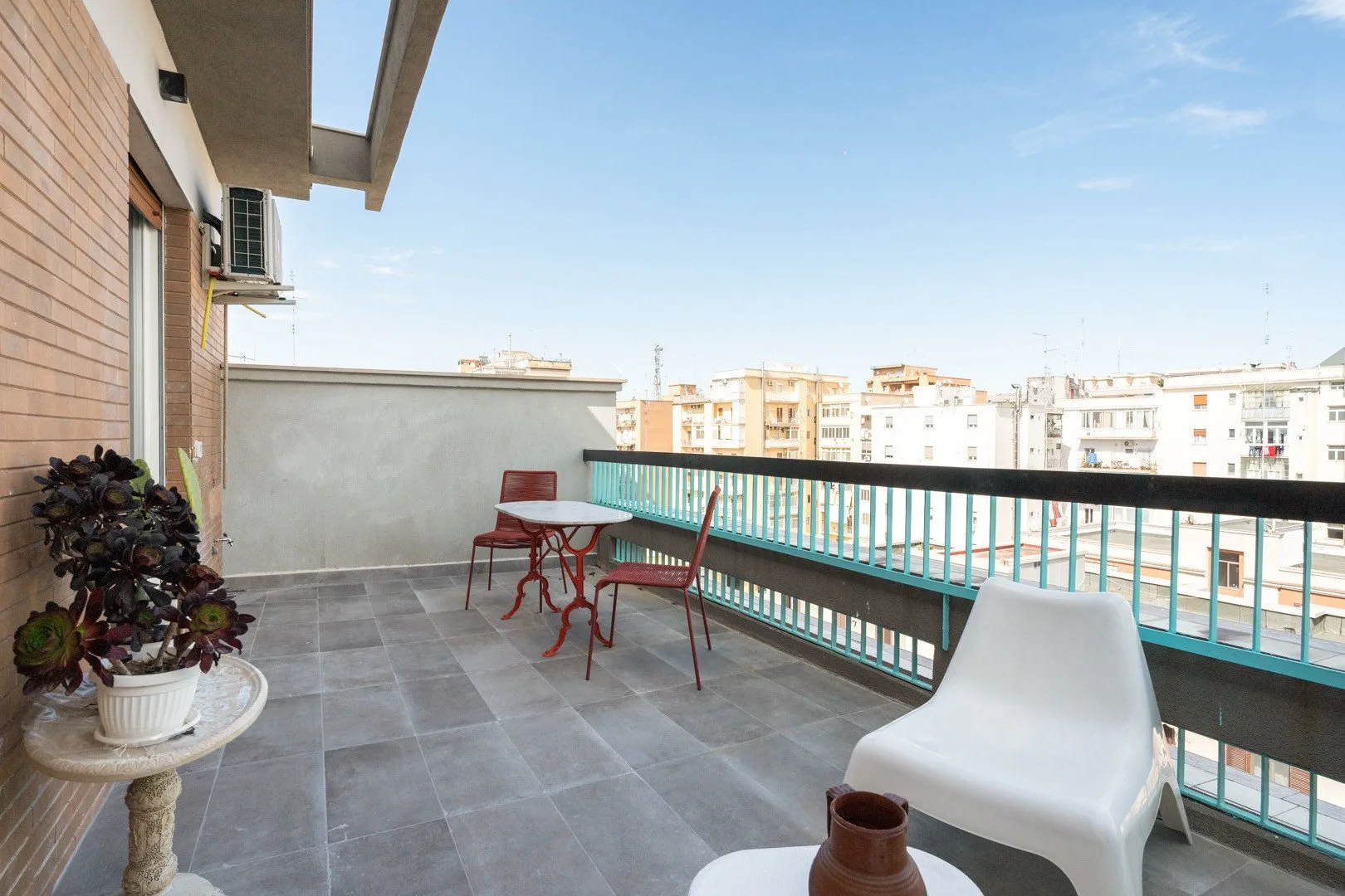 A tiled balcony with outdoor seating and a railing overlooking nearby buildings.