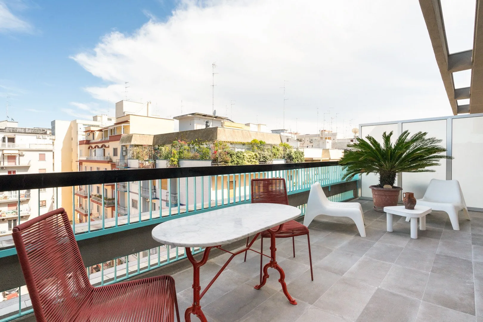 A furnished balcony overlooks nearby buildings, with a table, seating, and a potted palm.