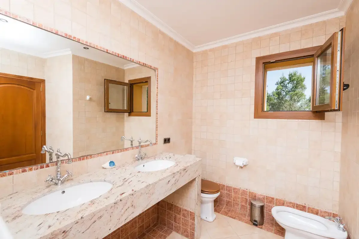 A tiled bathroom with a long counter, two sinks, and an open window.