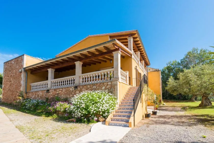 A yellow house has a covered porch, exterior stairs, and flowering shrubs.