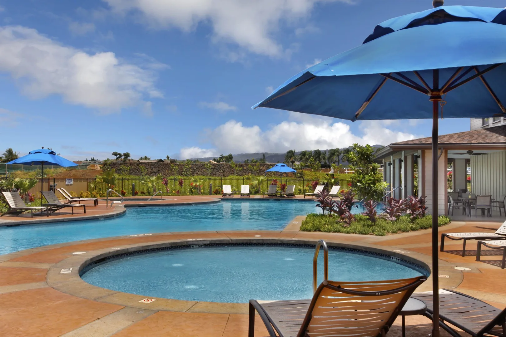 Una piscina exterior y un spa circular están junto a un patio con sombrillas azules.