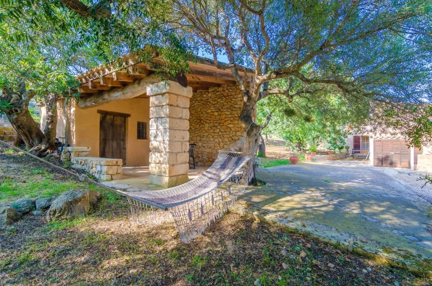 A stone cottage sits beneath leafy trees, with a hammock in the foreground.