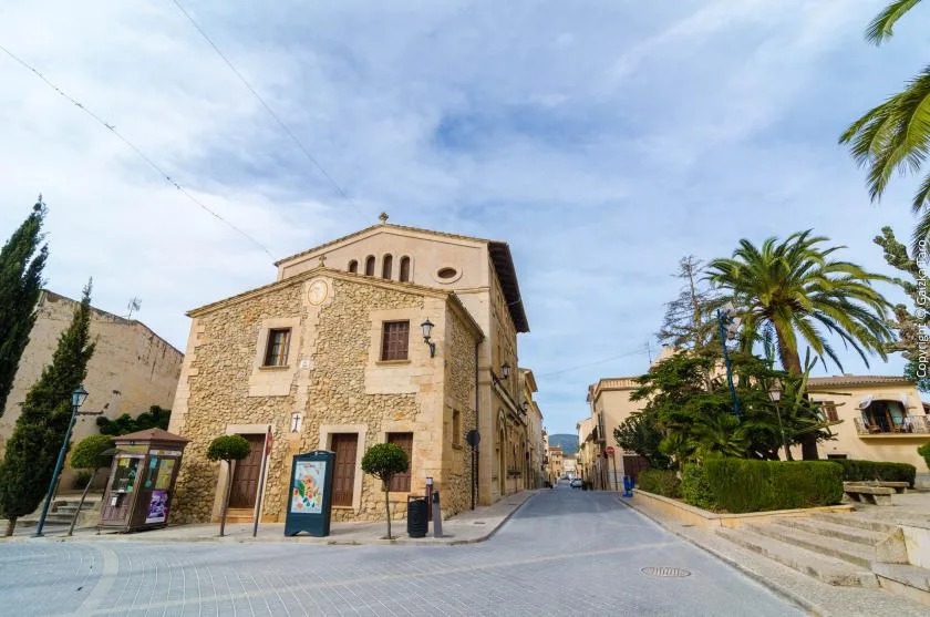 A paved street passes a stone building, with palm trees along the sidewalk.