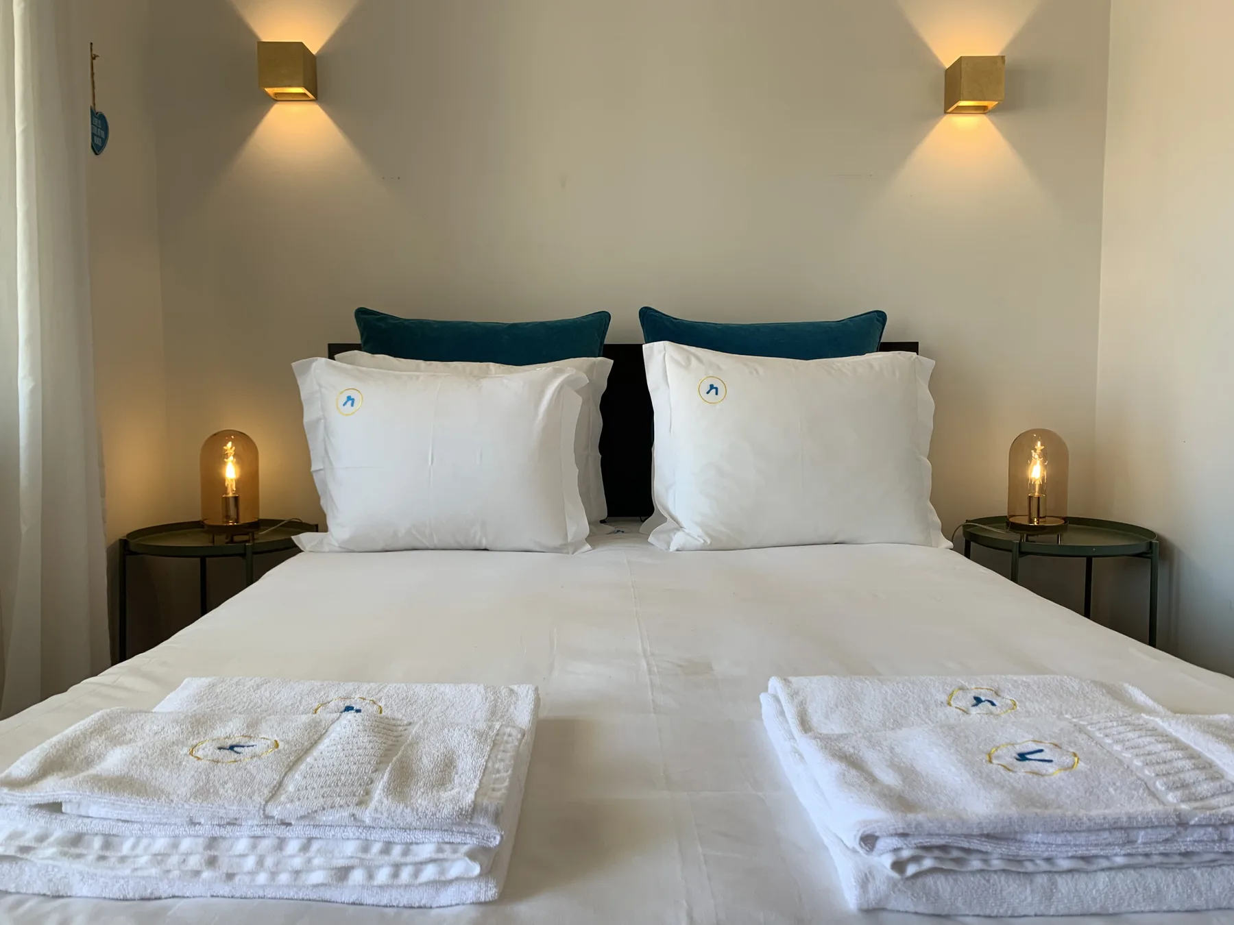 A bedroom with a neatly made bed, folded towels, and warm wall lighting.