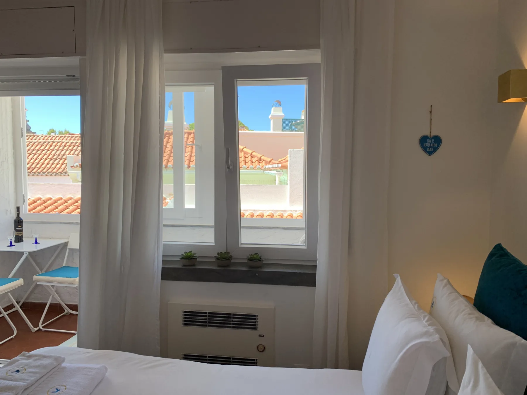 A bedroom with a bed beside bright windows overlooking nearby rooftops.