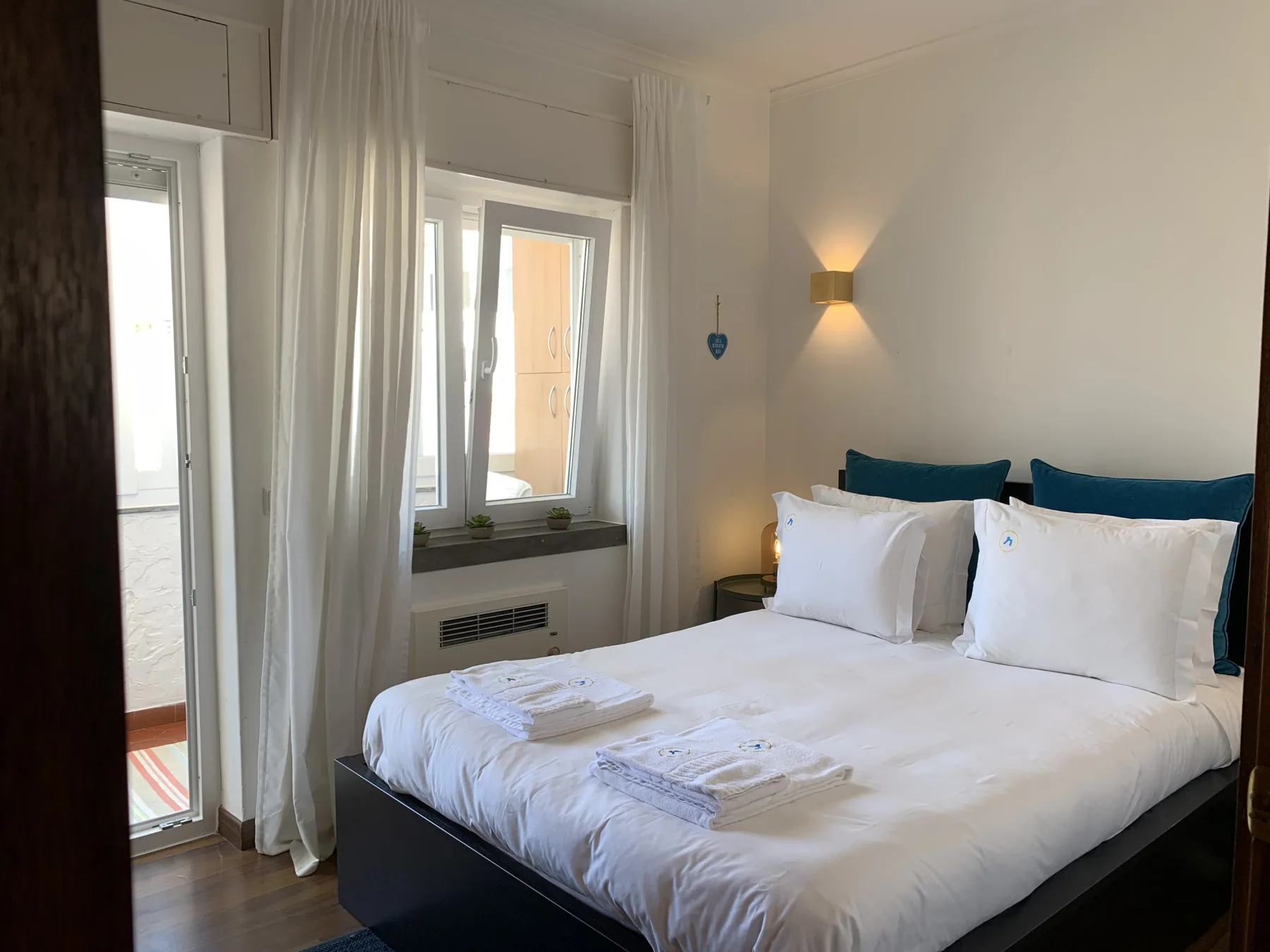 Dormitorio con una cama, ropa de cama blanca y ventanas altas con cortinas.