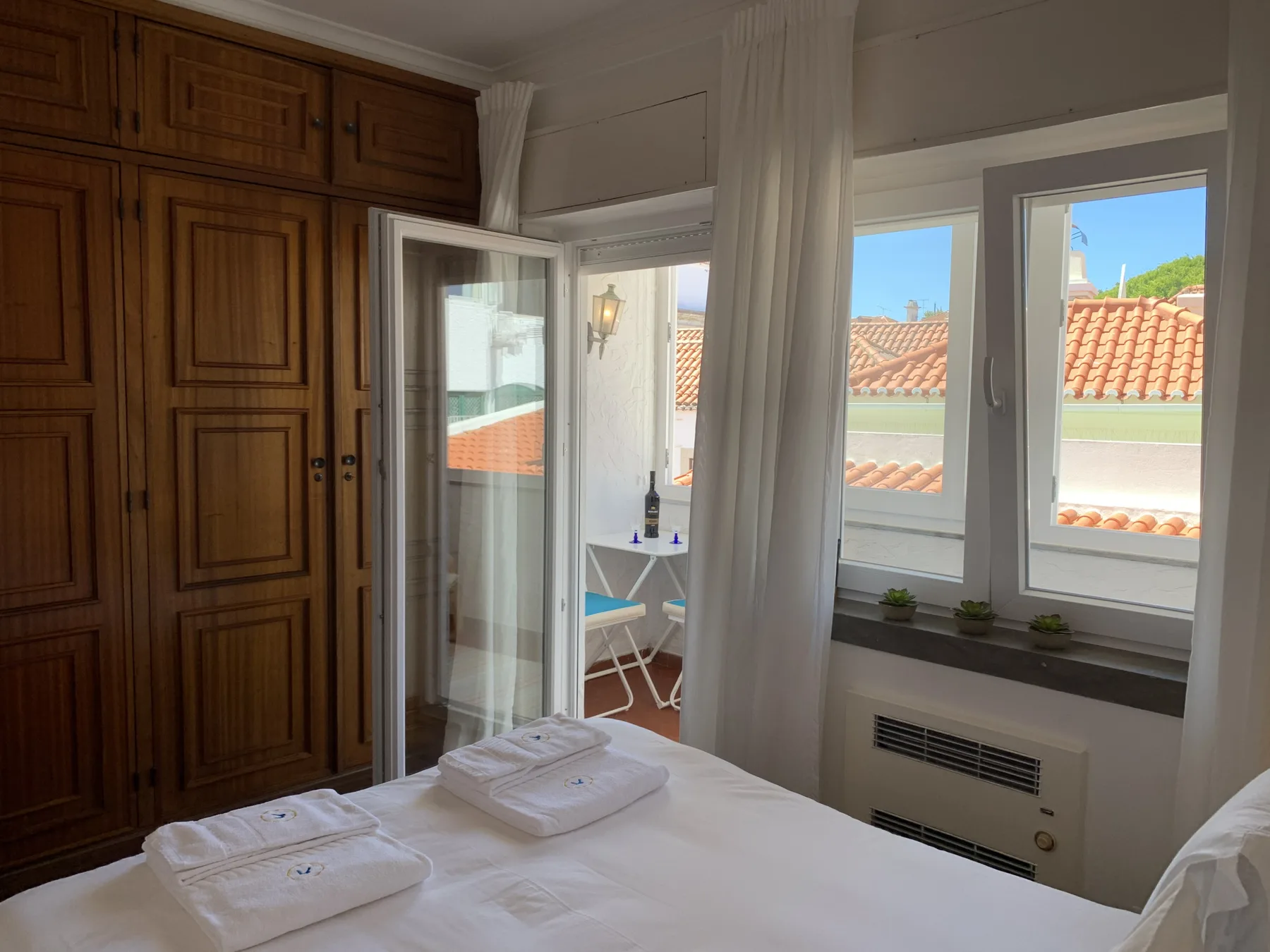 A bedroom with a bed, wooden wardrobes, and windows overlooking tiled rooftops.