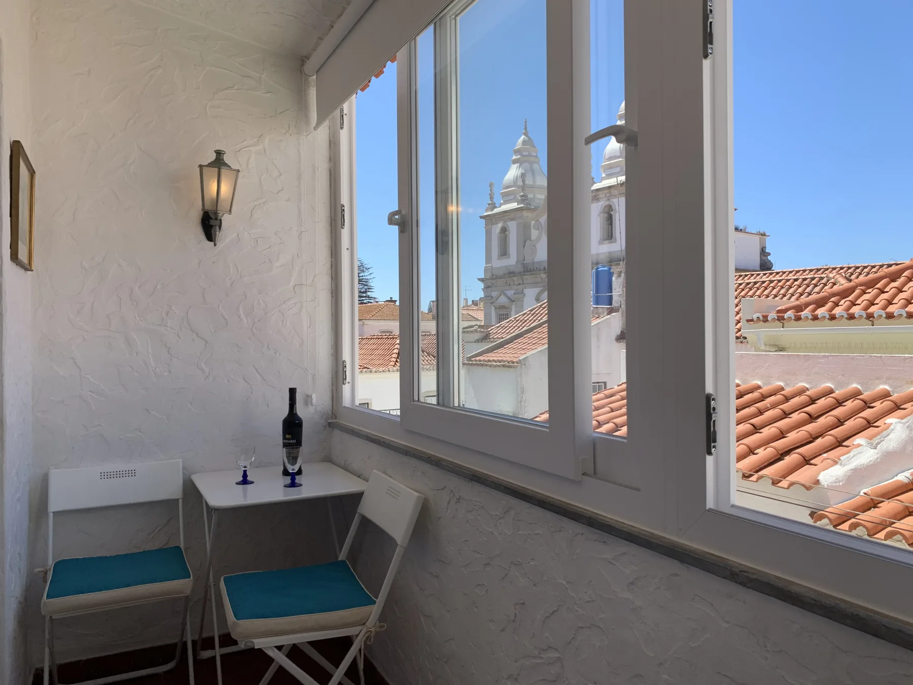 A bright sitting area beside large windows overlooks tiled rooftops and a church tower.