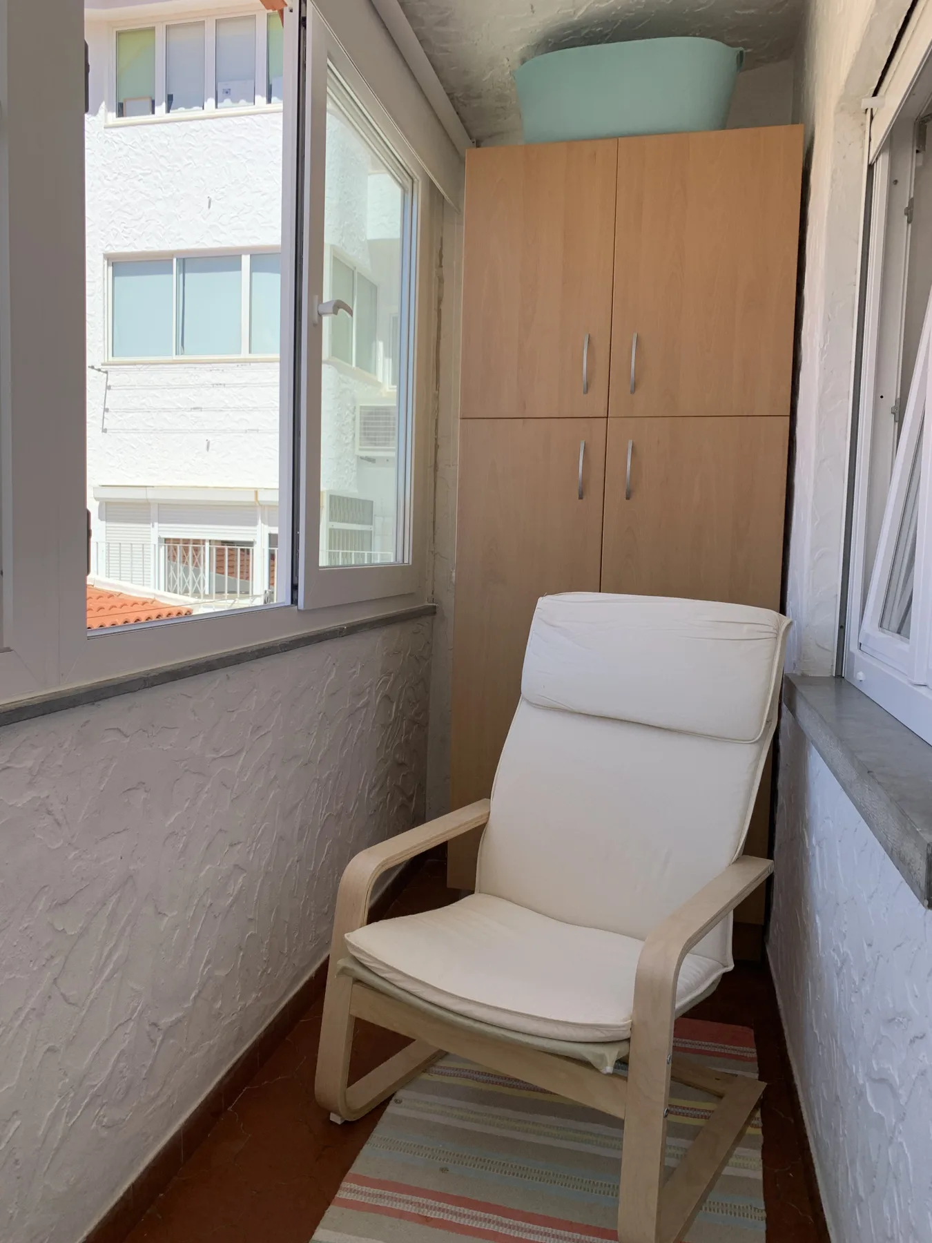 A narrow enclosed balcony has a cushioned chair beside a wooden cabinet.