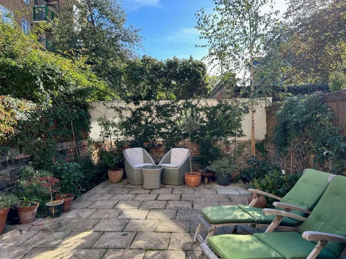 Un patio ajardinado pavimentado con asientos, plantas en macetas y vegetación alrededor.