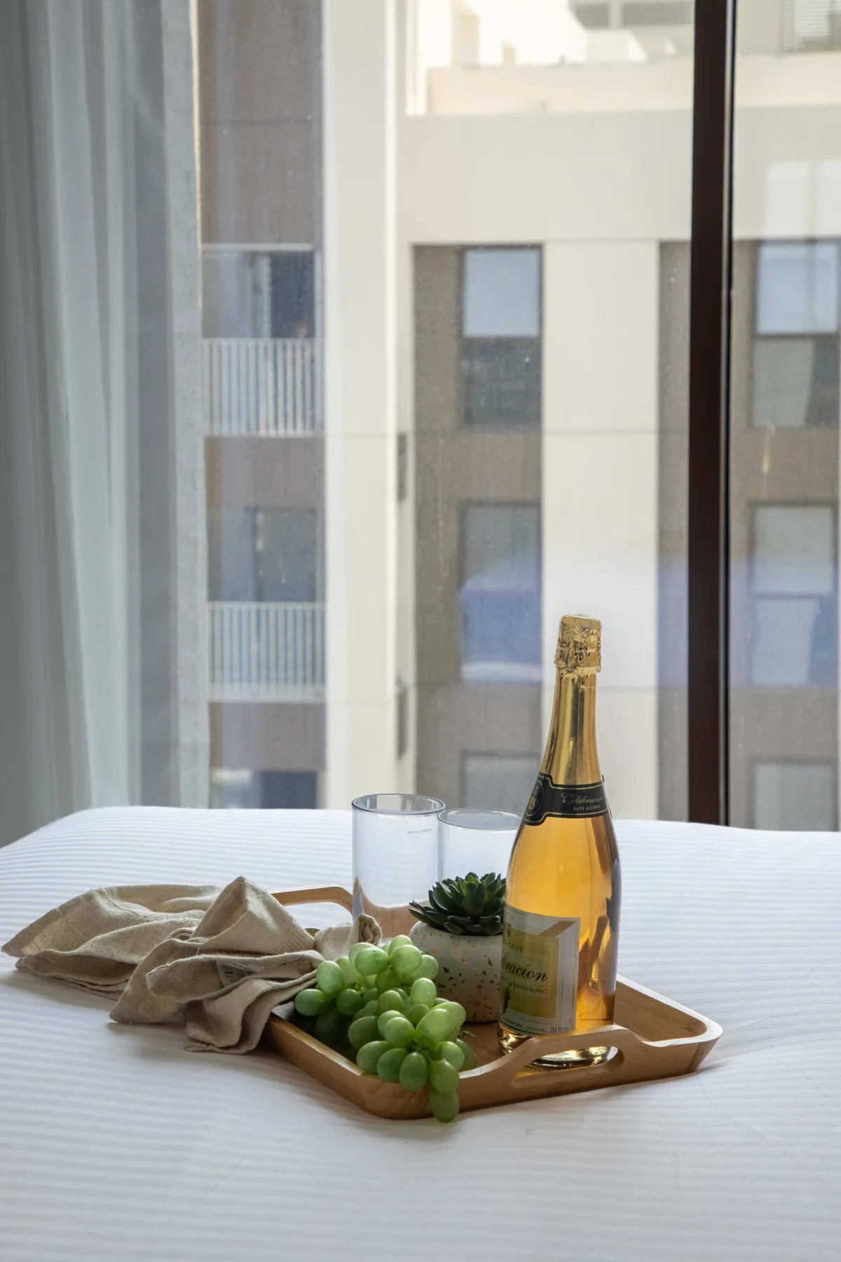 Una cama junto a una ventana grande, con una bandeja con fruta y una botella.