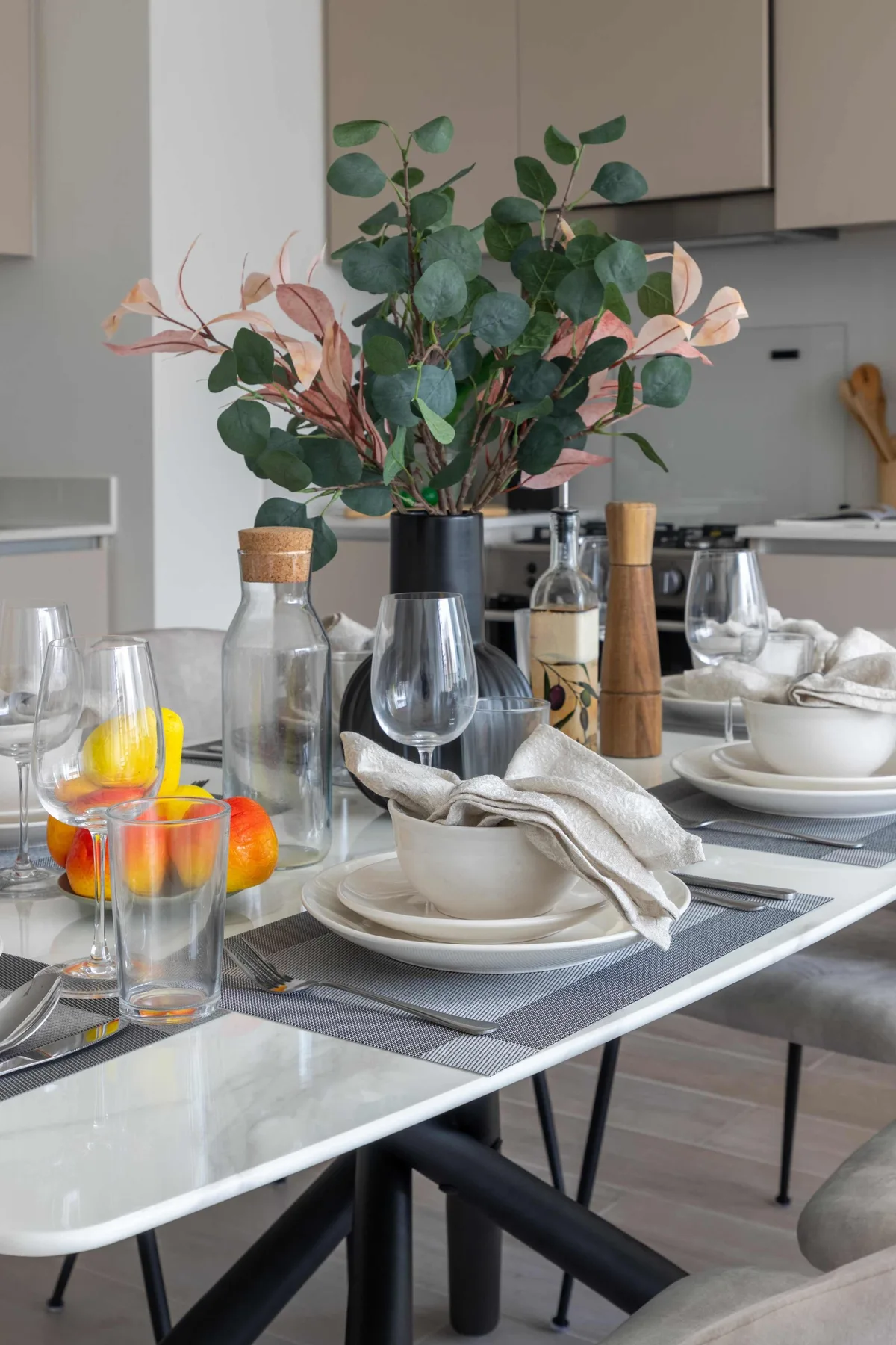 Una mesa de comedor está puesta con platos y vasos junto a un centro de mesa con hojas.