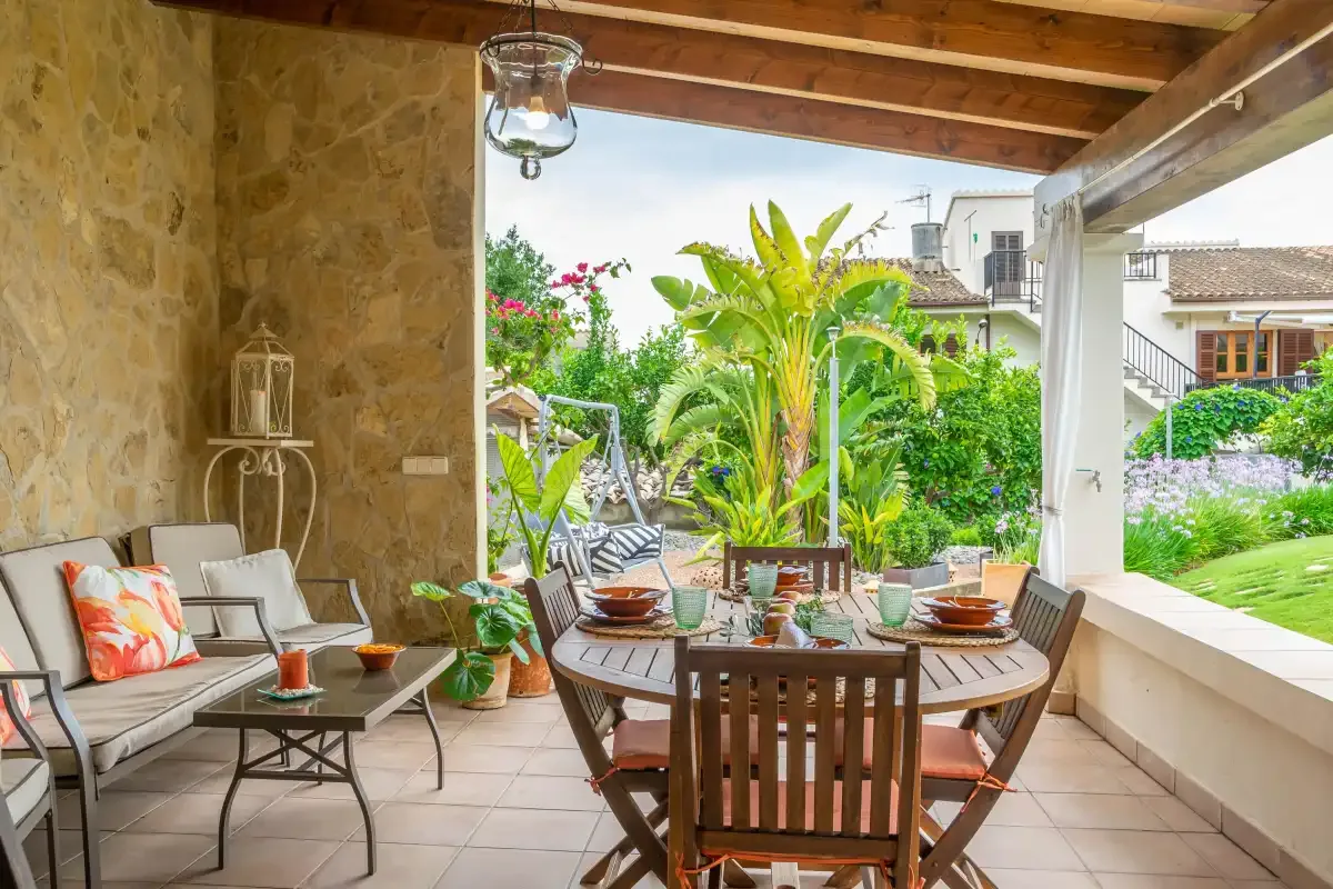 Un patio cubierto tiene una mesa de comedor y asientos junto a un jardín frondoso.