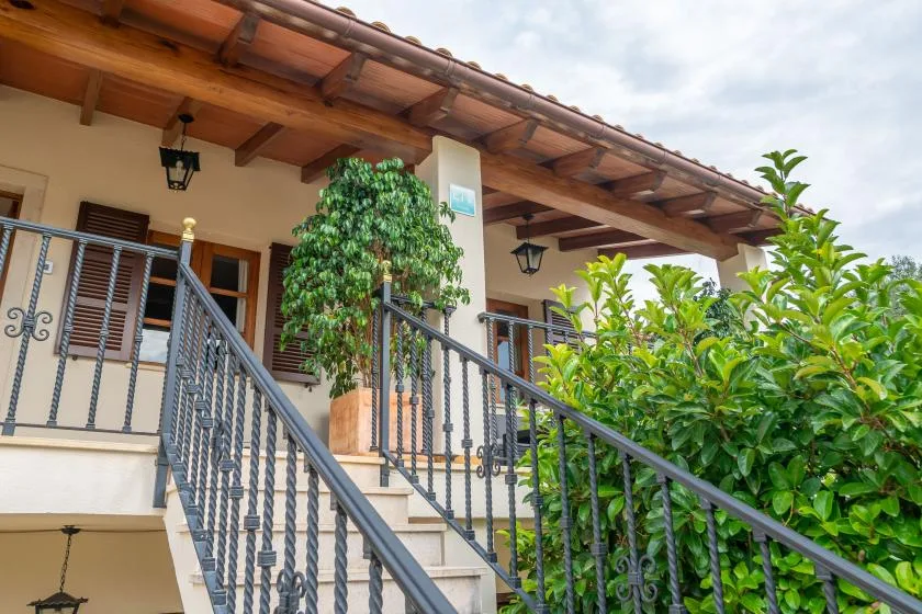 Una escalera exterior con barandillas metálicas conduce a un balcón cubierto junto a plantas frondosas.