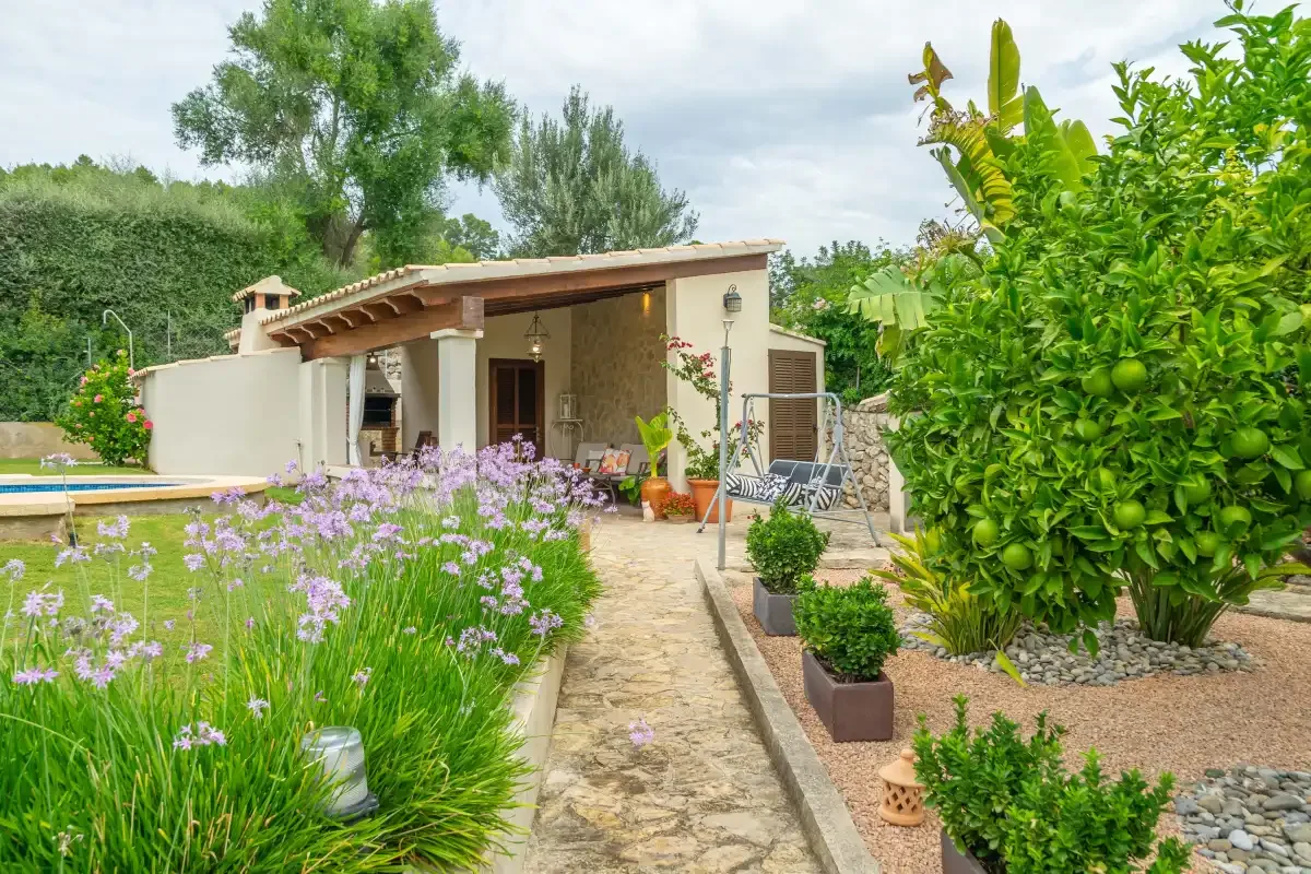 Un sendero del jardín conduce a un patio cubierto junto a plantas en flor y una piscina.