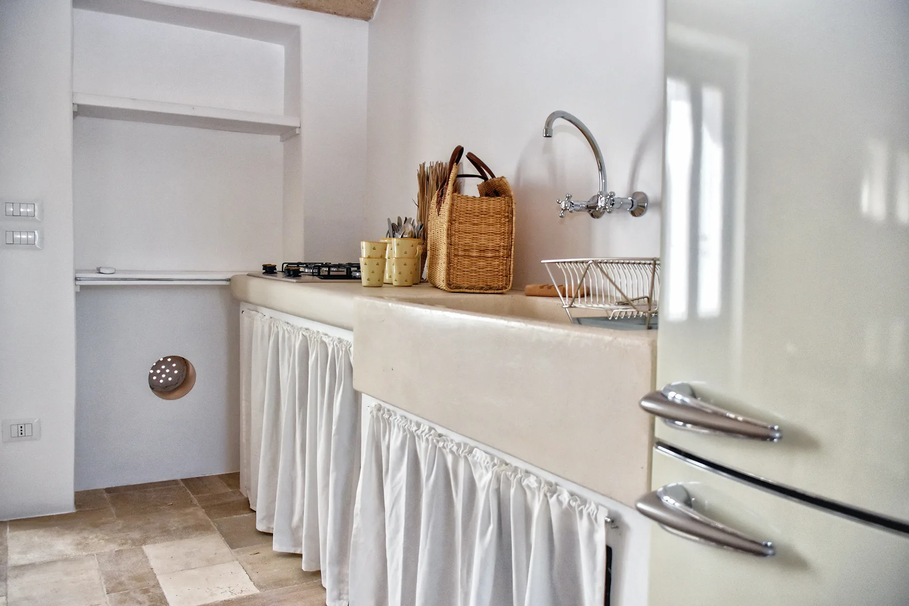 Una pequeña cocina luminosa con encimera, grifo de pared y almacenamiento con cortinas.