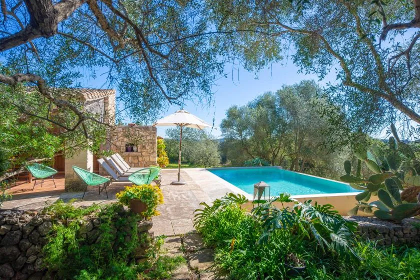 An outdoor swimming pool beside a stone patio with lounge seating and an umbrella.